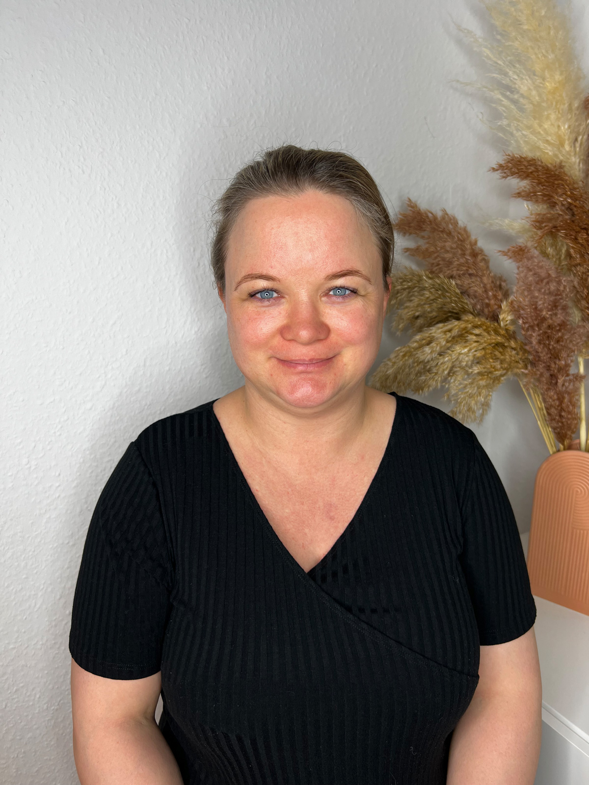 Ein frontales Portrait von mir selbst vor einer weißen Wand mit beigen Farn rechts hinter dem Modell.